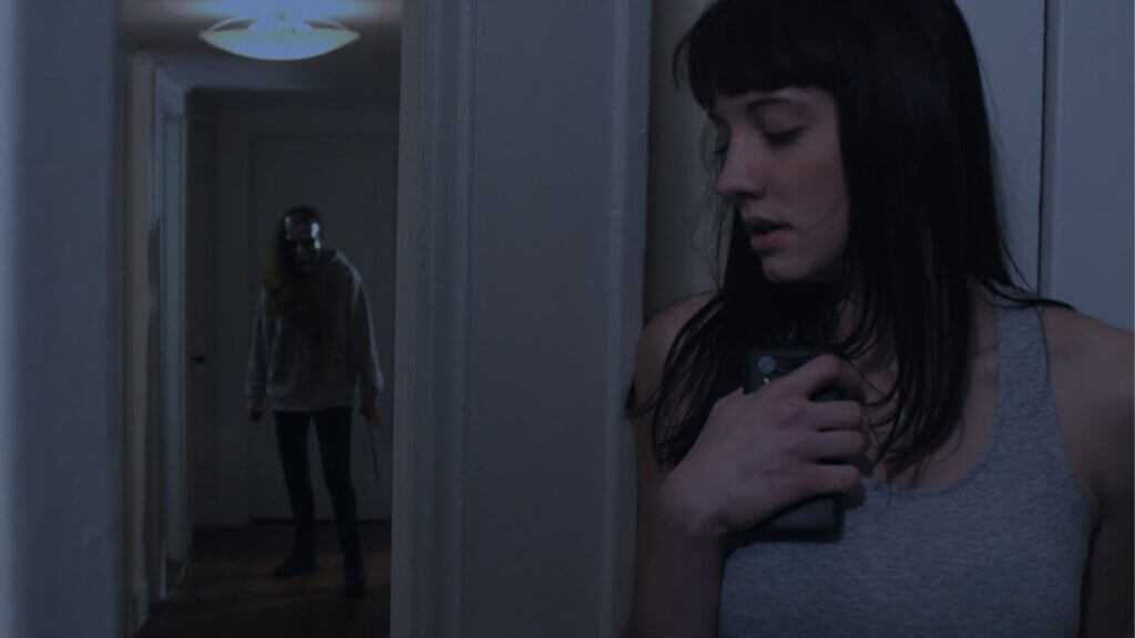 a scared girl in grey top holding her phone while a masked man with a knife stands behind her in the hallway